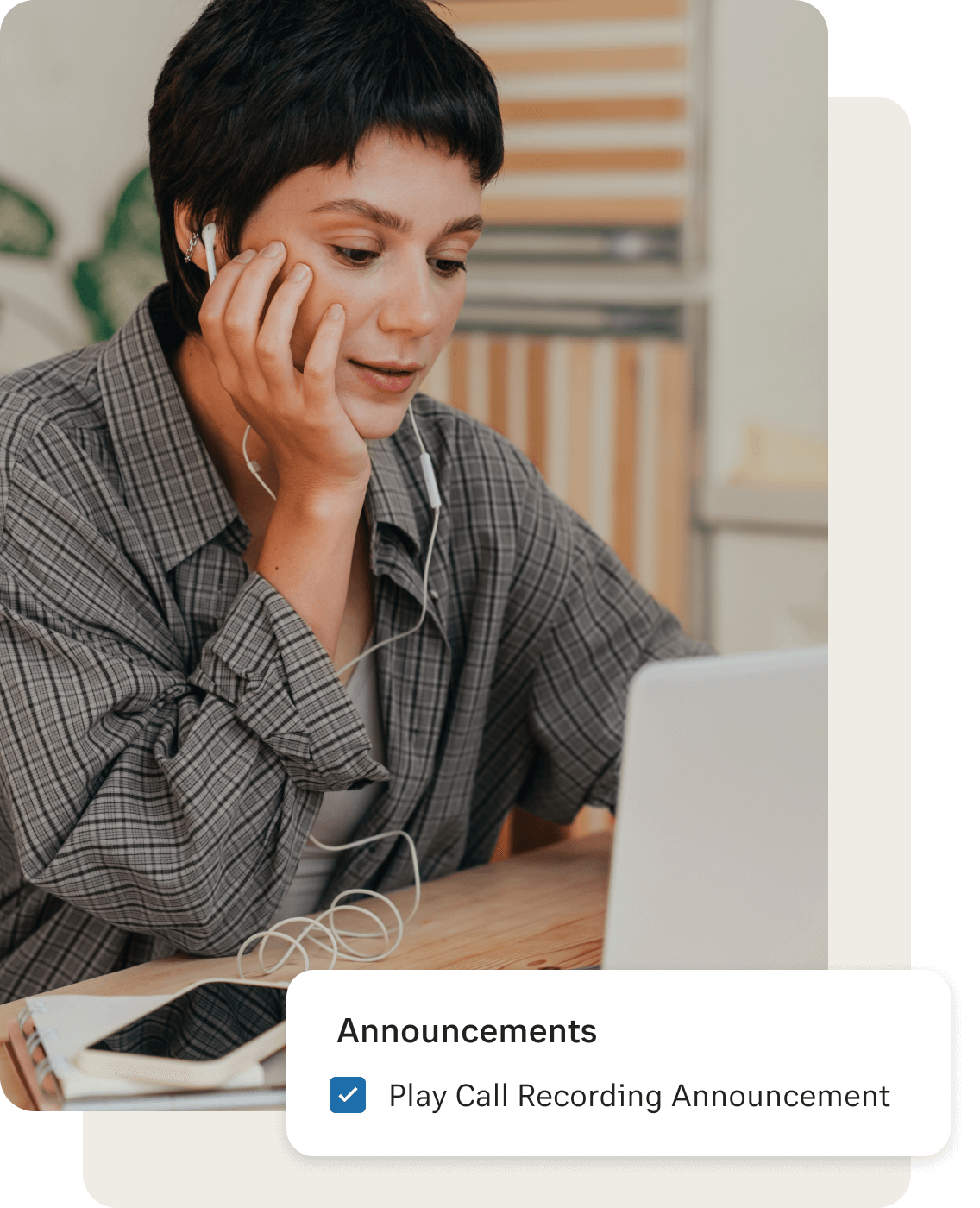 A woman with earphones connected to her mobile phone while looking at her laptop is listening to the call recording announcement
